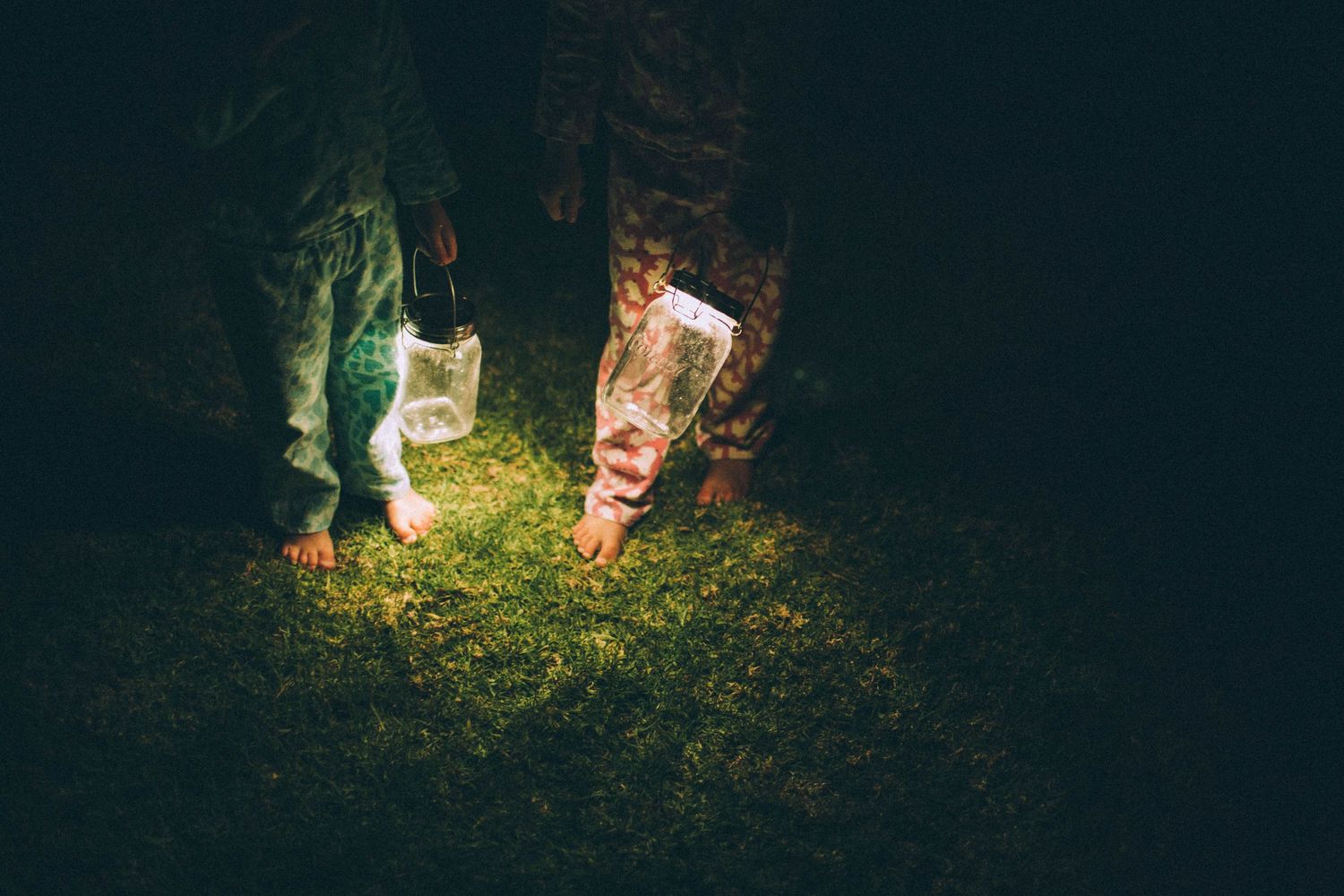 Two barefoot children stand on glowing green grass at night holding clear plastic bags.