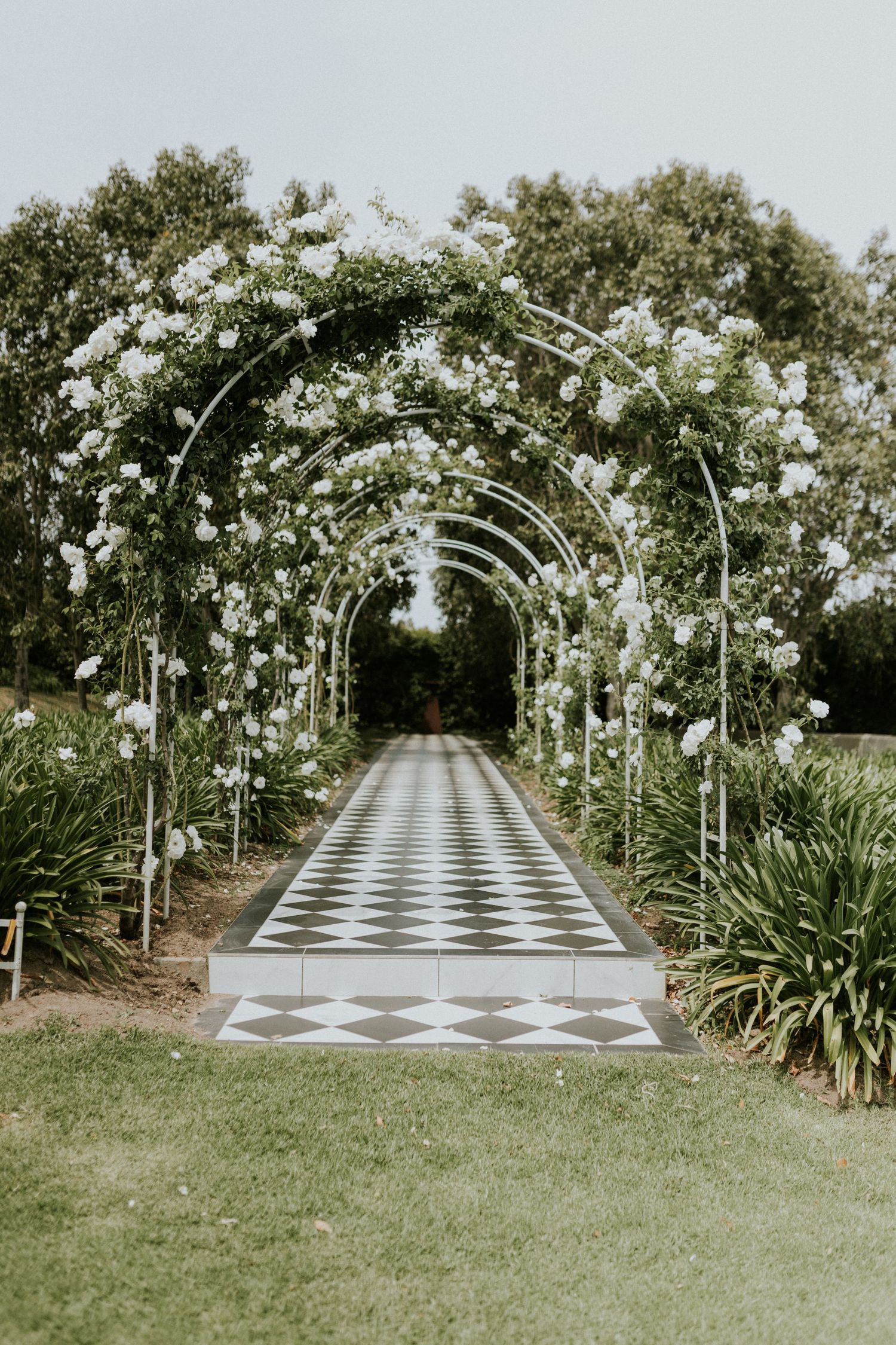 Beautiful garden wedding aisle with white floral arches over a black and white checkered path.