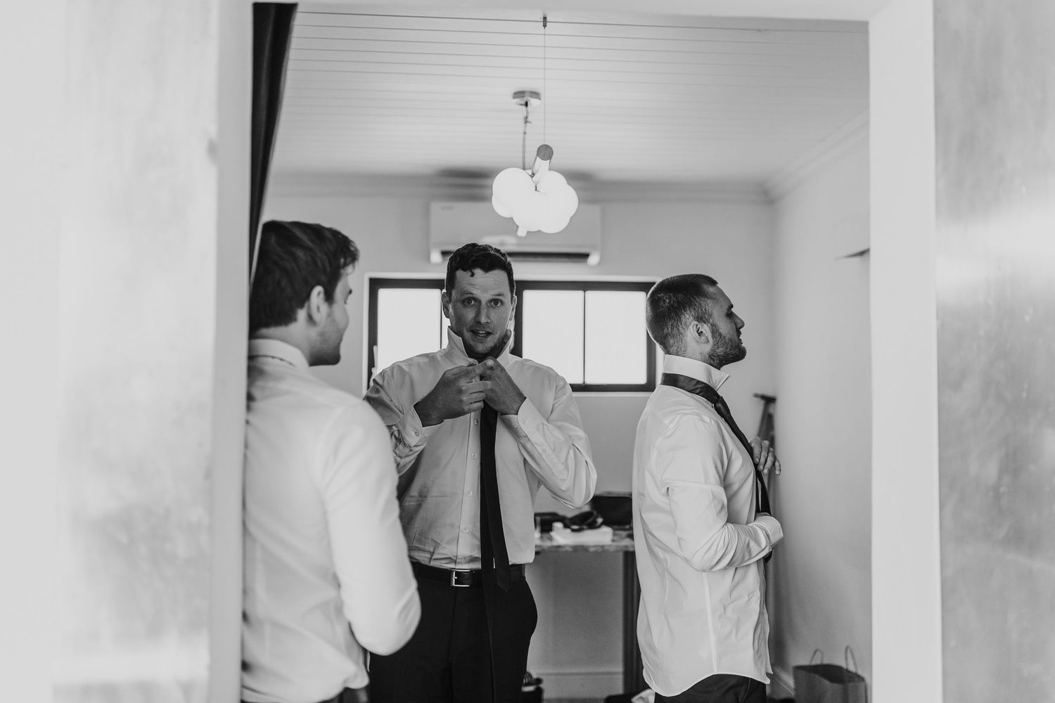 Black and white photo of groomsmen helping each other with ties and suspenders while getting ready.