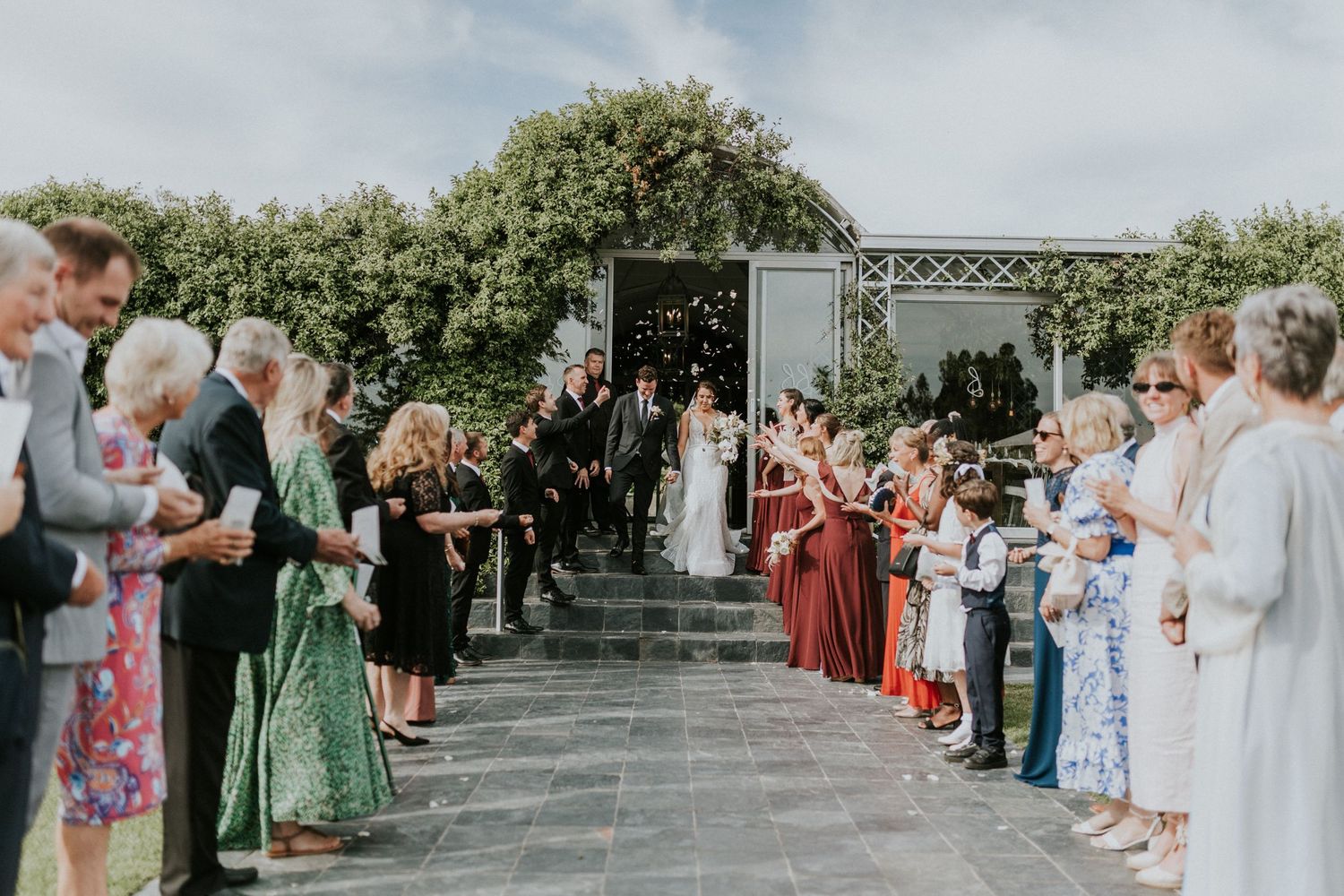 Wedding guests line up to celebrate the newlyweds as they walk through a confetti-filled exit at an outdoor ceremony.