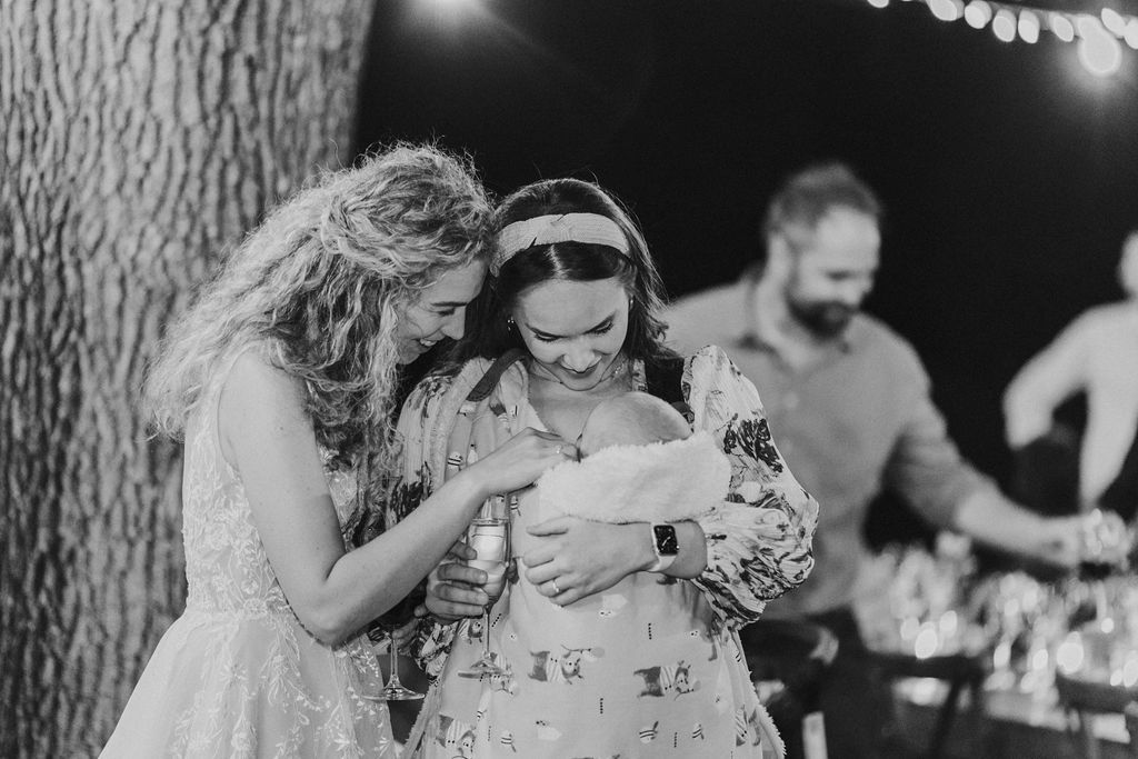 Black and white photo of wedding guests admiring a newborn baby under string lights at reception.