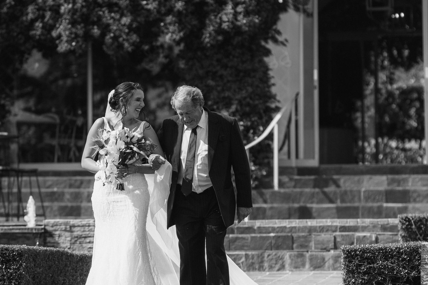 Black and white photo of a bride in a veil smiling at her father as they walk down steps toward the ceremony.