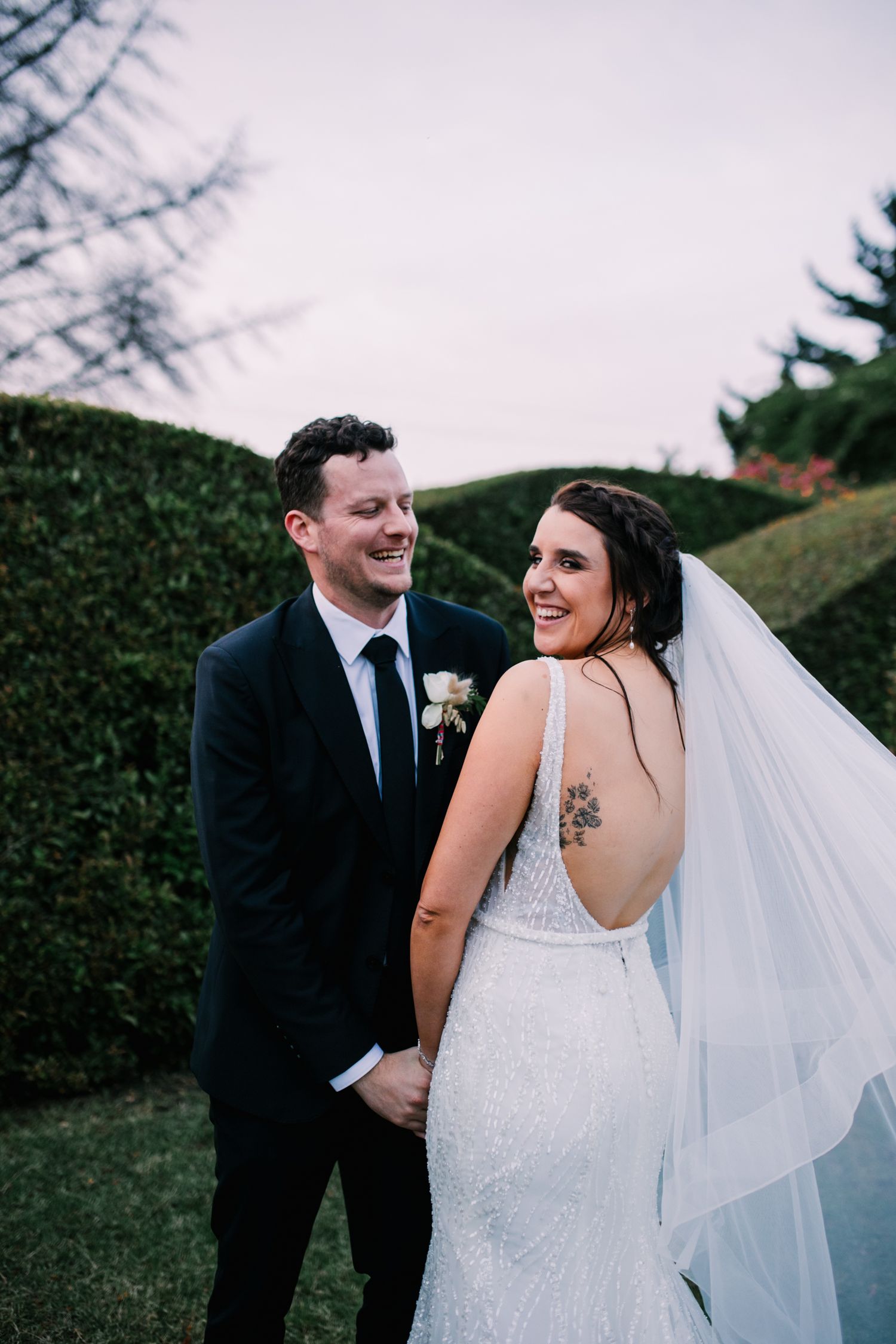 Smiling bride and groom holding hands in front of tall green hedges at golden hour.