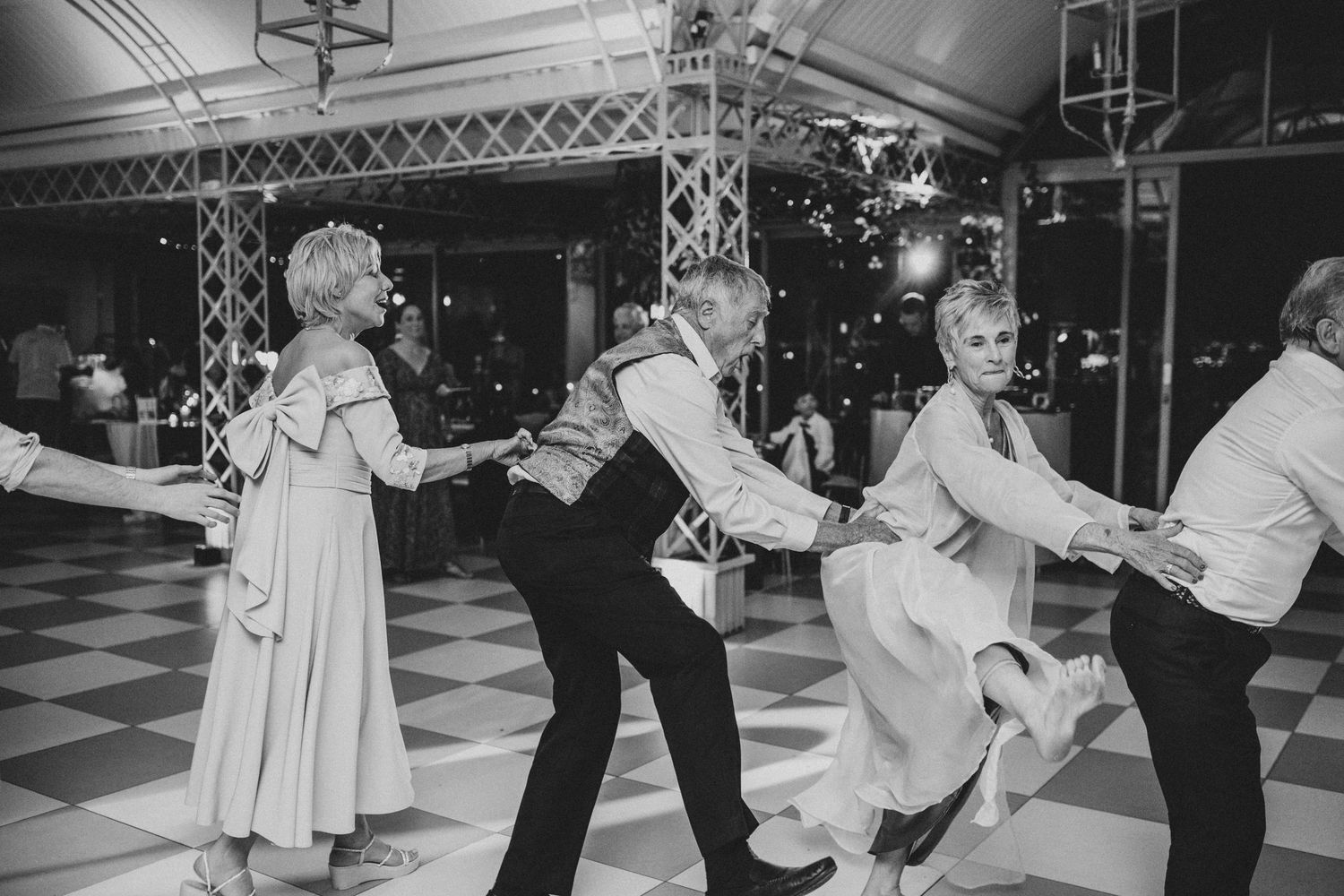 Elderly wedding guests joyfully holding hands and dancing together on a checkered ballroom dance floor.