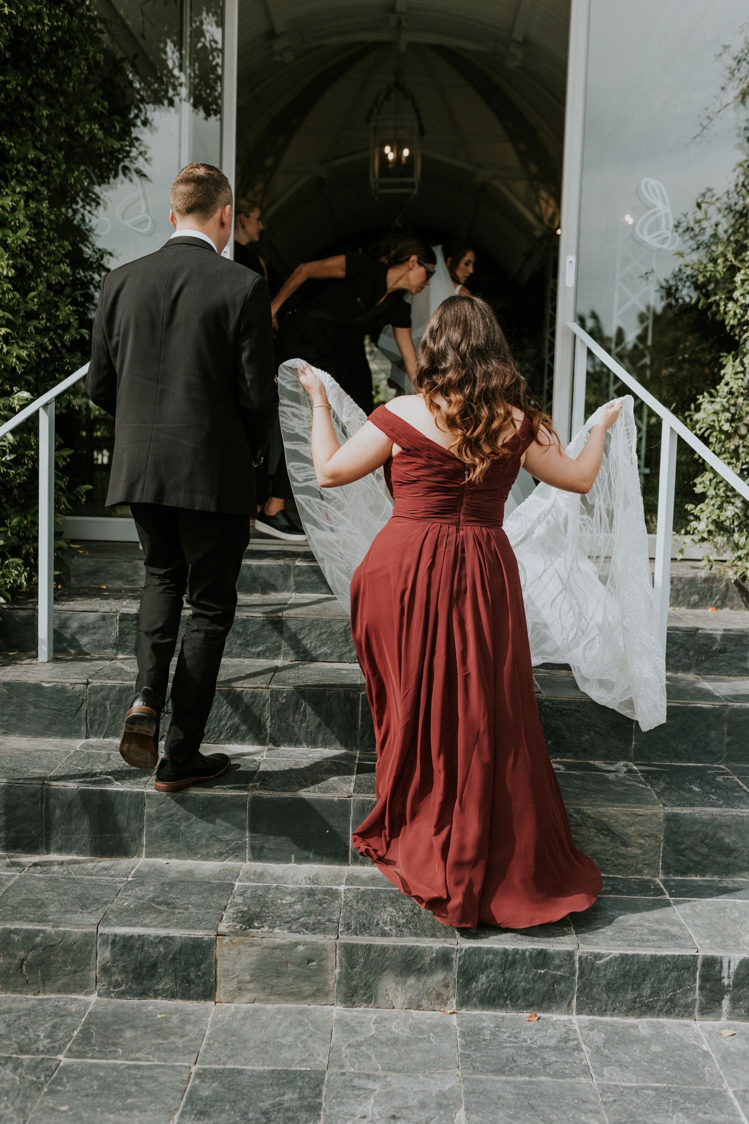 Bridesmaid in red gown carries the bride's long white veil up marble steps toward a white wedding venue.