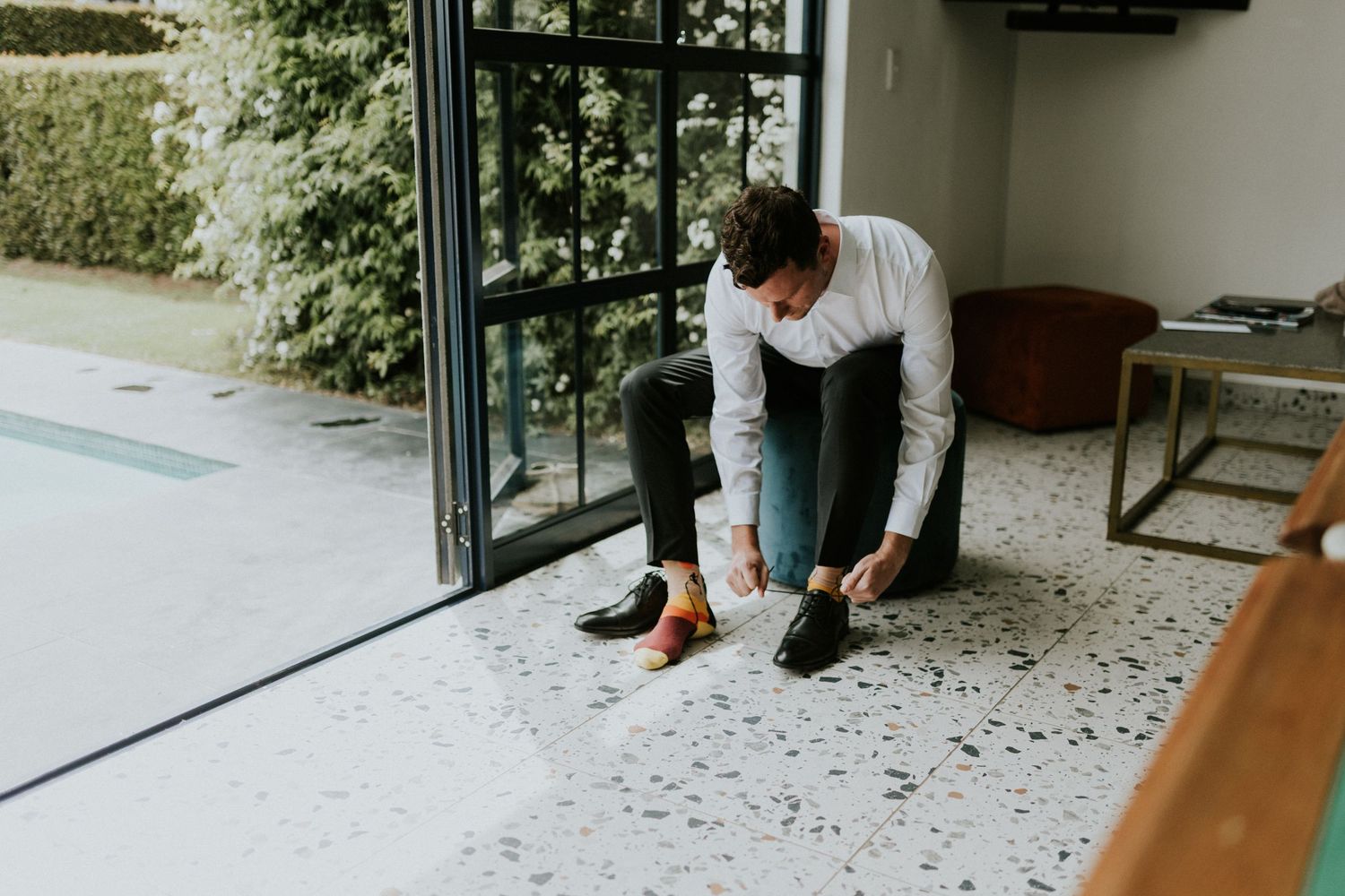 Groom tying his dress shoes on terrazzo floor beside glass doors overlooking a garden pool.