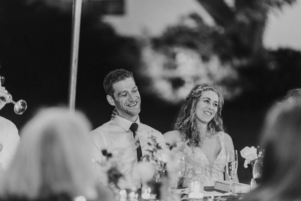 Happy bride and groom smiling while listening to wedding speeches at outdoor reception dinner.
