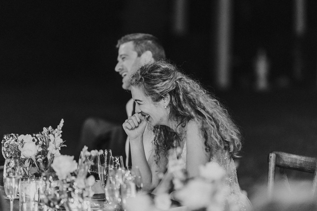 Emotional bride covers mouth while laughing at wedding reception dinner table speech.