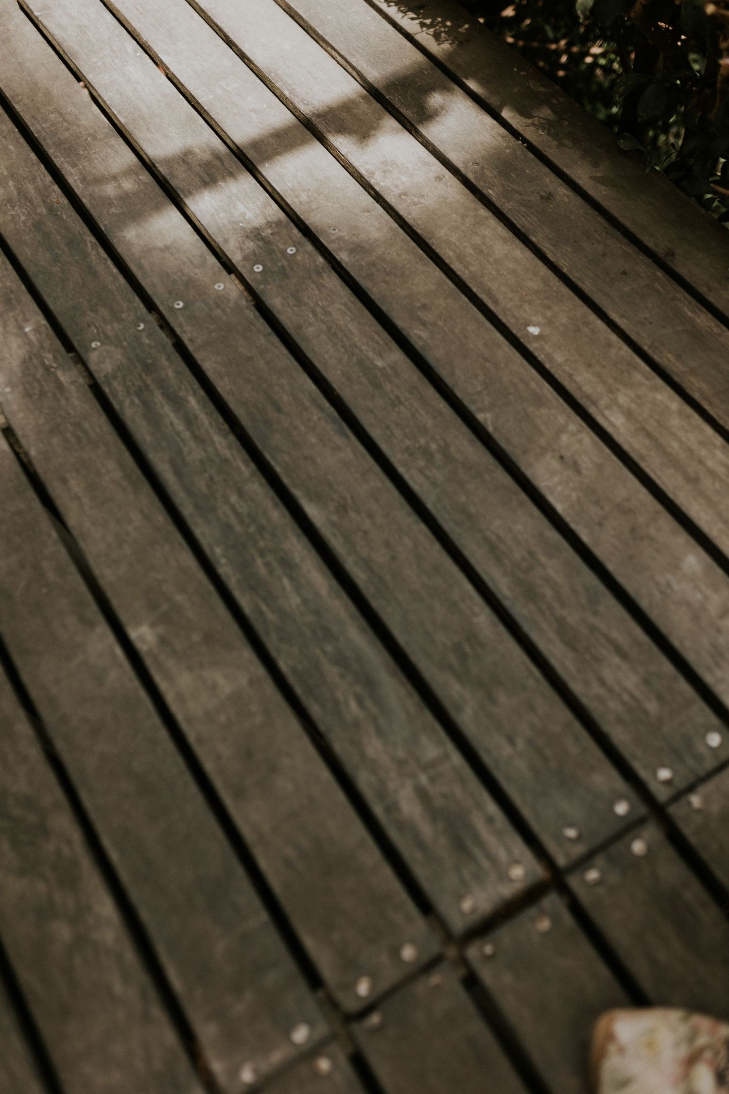 Close-up detail of weathered gray wooden deck planks with natural texture and subtle light shadows from nearby greenery.
