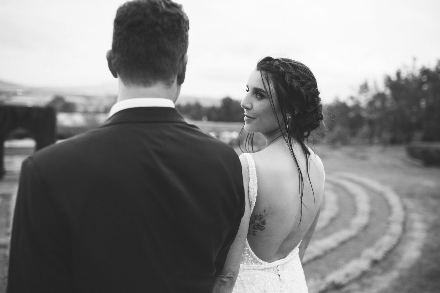 Black and white photo of bride with back tattoo in open-back wedding gown gazing at her groom.