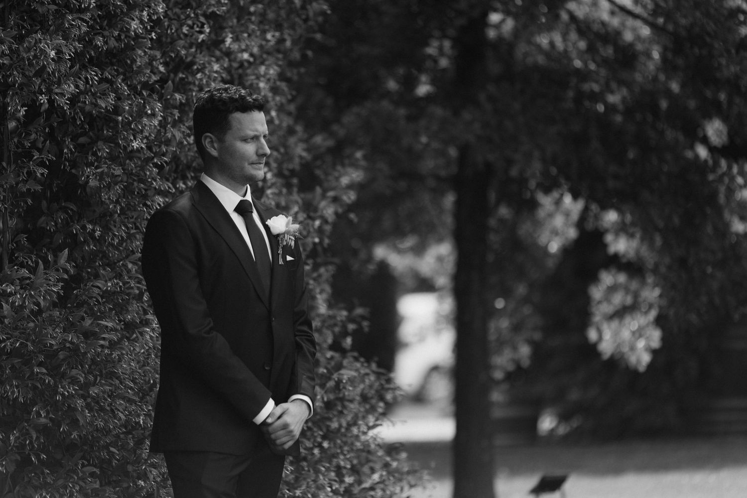 Black and white portrait of a handsome groom in a dark suit waiting among lush green hedges before the ceremony.