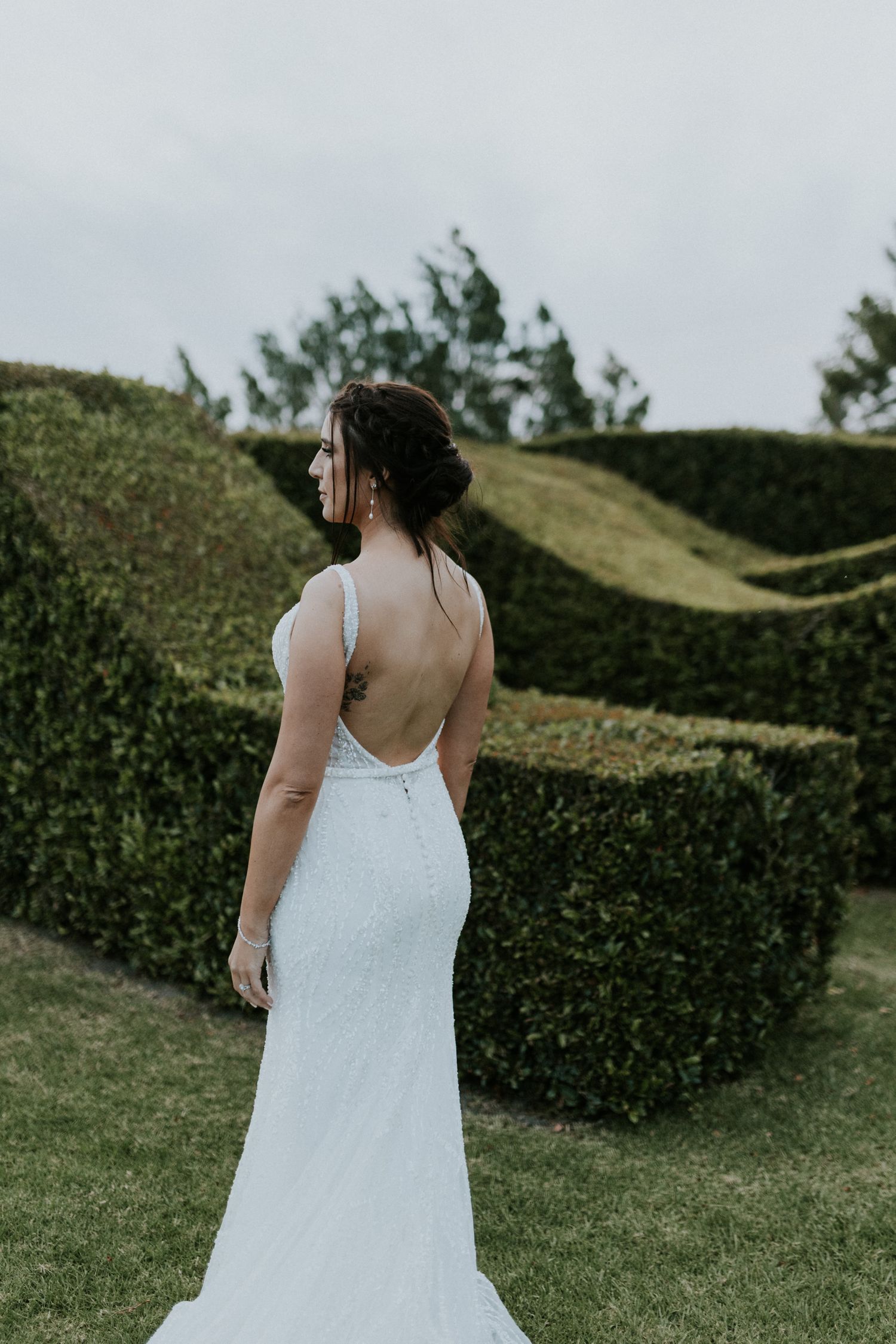 Bride in elegant backless white wedding gown with low back detail standing before manicured hedges.