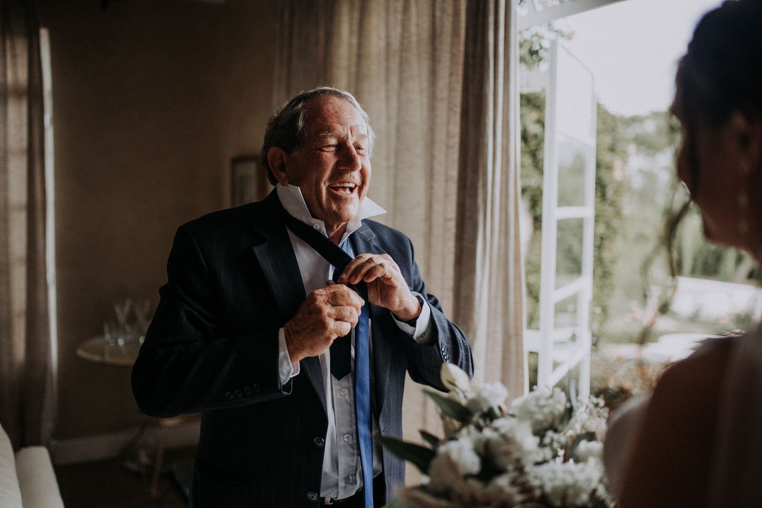 Smiling father of the bride adjusting his blue bow tie while holding white flowers.