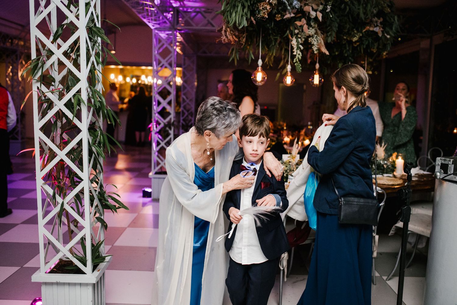 Woman adjusting a young boy's tie at a wedding reception with purple lighting and hanging greenery decor.