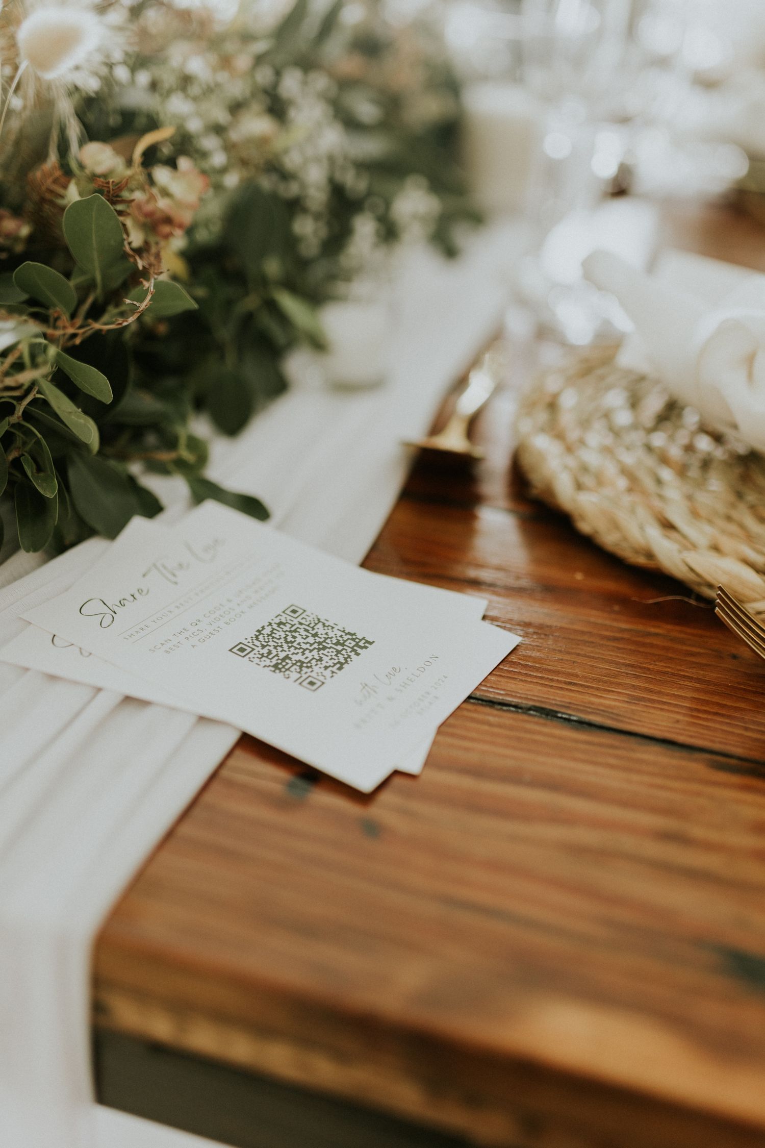 Rustic wooden wedding reception table with QR code card, floral centerpiece, and white table runner.