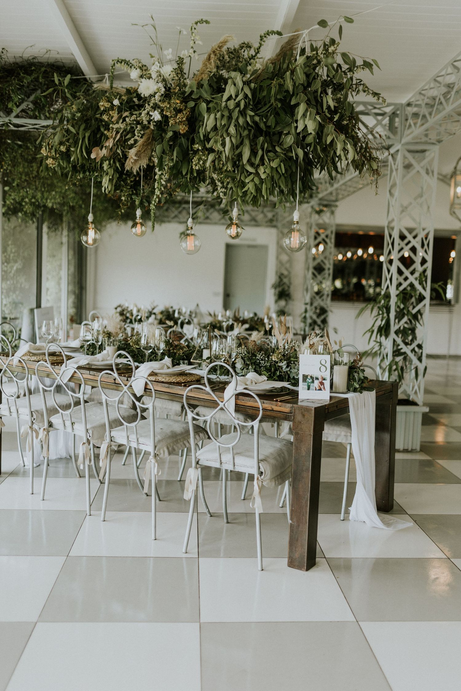 Elegant wedding reception hall with hanging greenery, Edison bulbs, and white iron chairs.