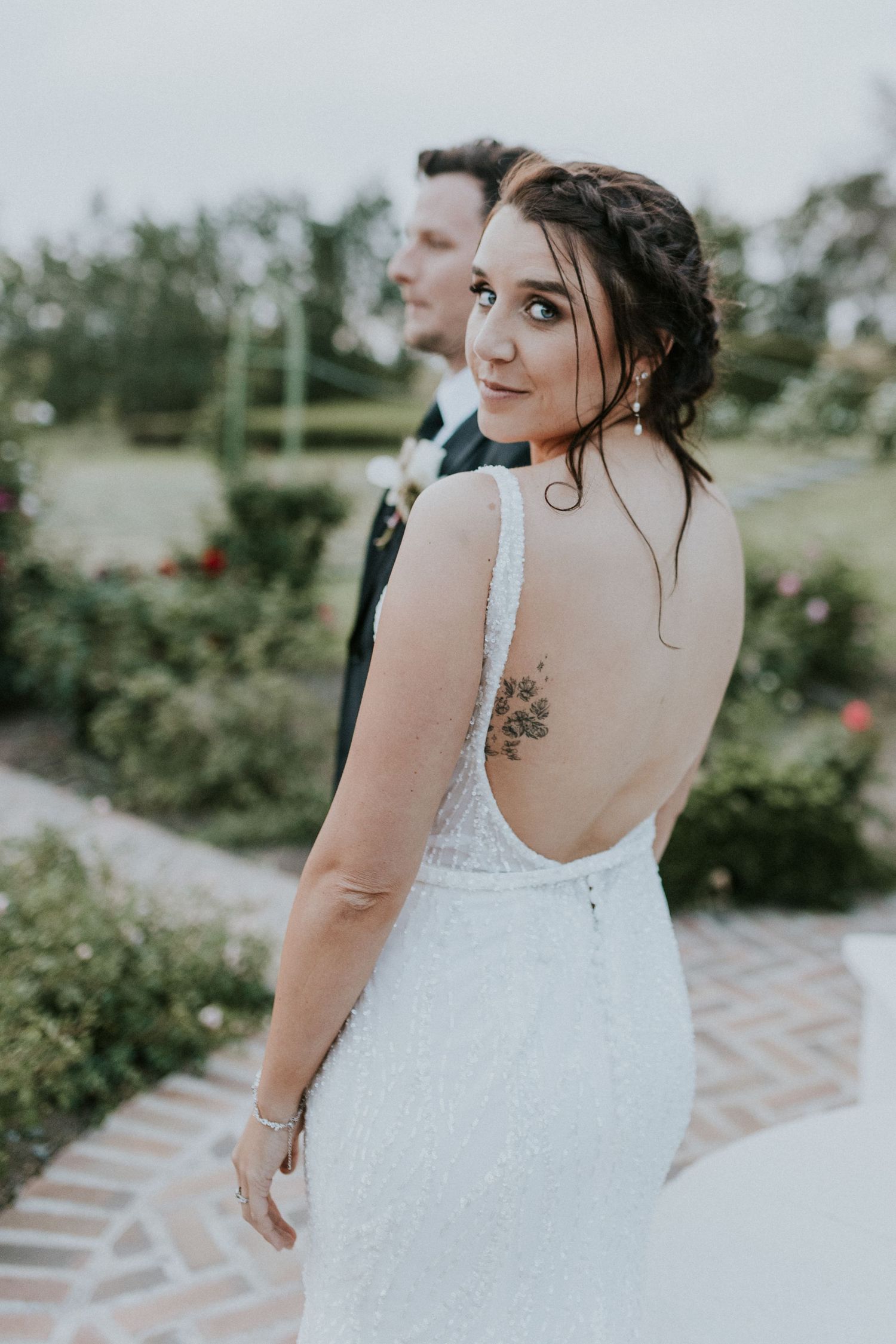 Bride in backless beaded wedding gown with floral tattoo glances back while groom stands in garden.