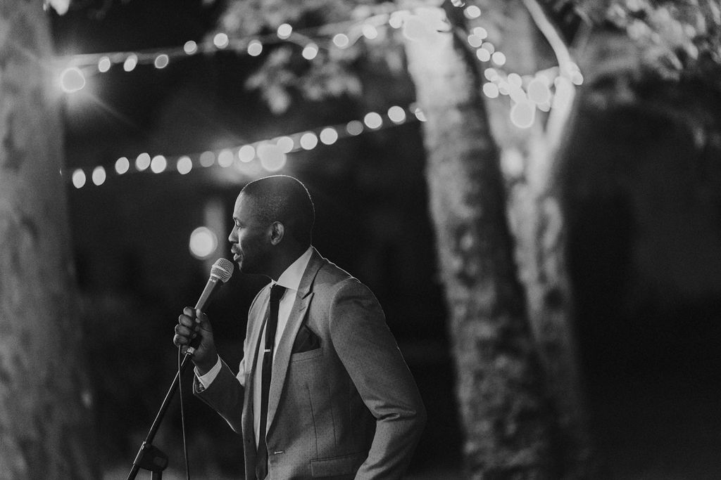 Elegant man in suit delivering heartfelt speech at microphone during outdoor evening wedding.