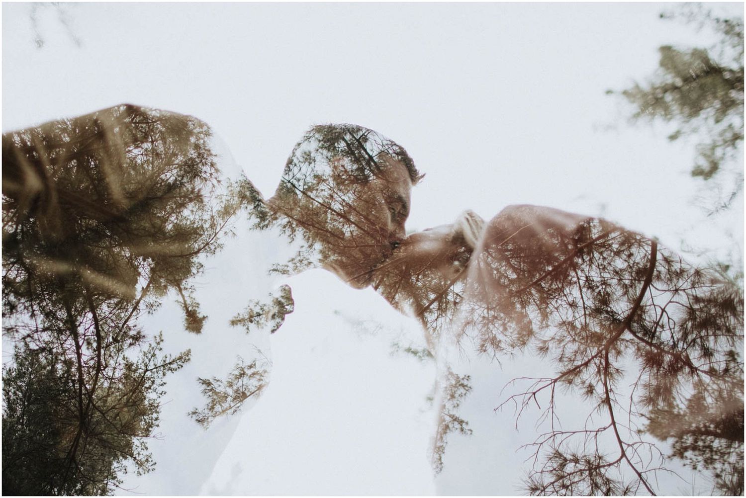 Dreamy double exposure photo of a couple kissing blended with tall forest trees against a bright sky.