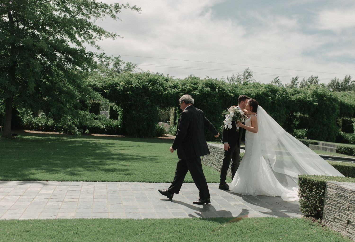 Bride in a long veil shares a kiss with her father while a groomsman walks ahead through a beautifully landscaped garden.