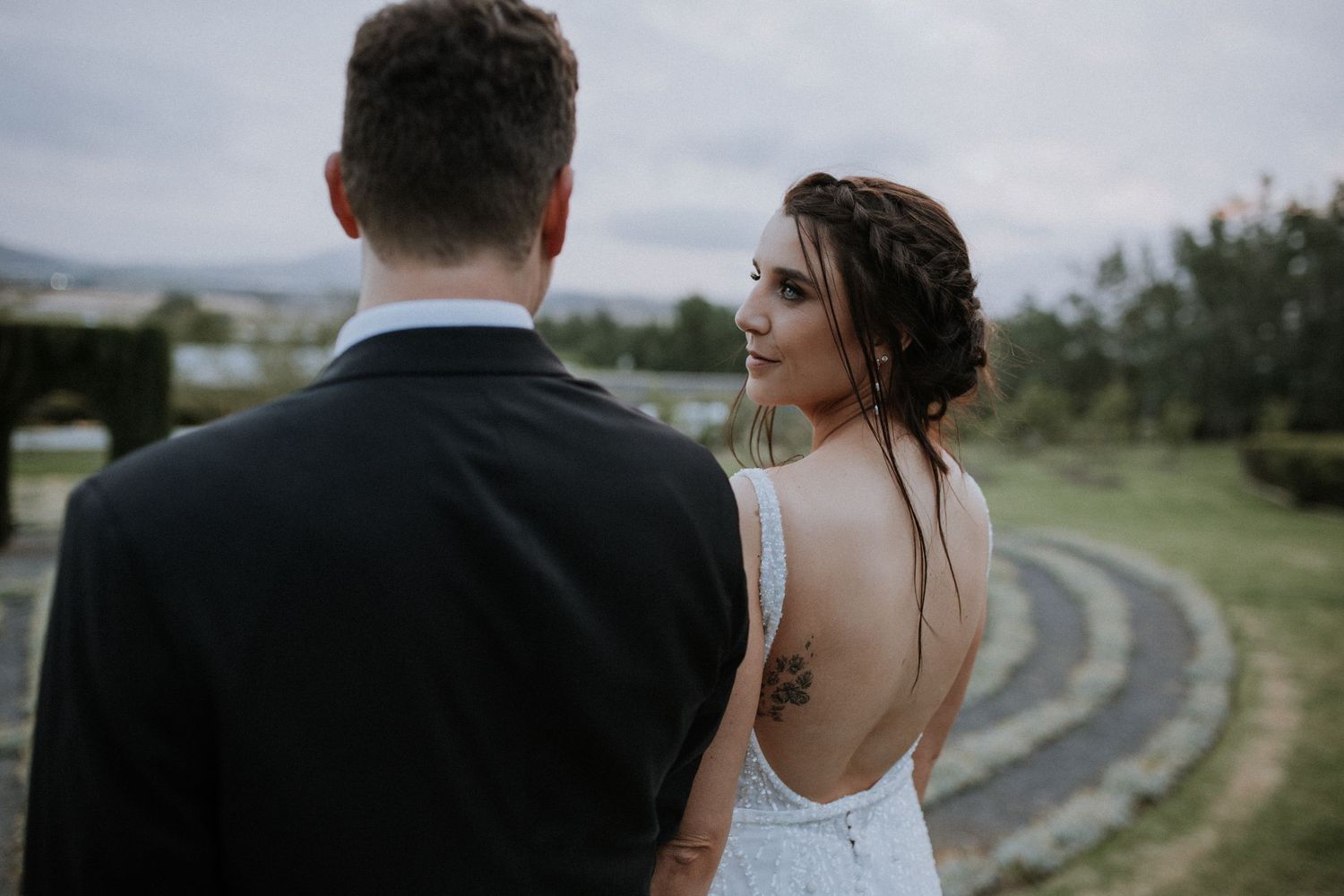 Bride in backless lace gown with tattoo looks over shoulder at groom in black suit at outdoor wedding.