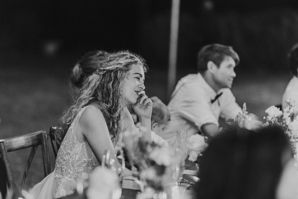 Bride laughs joyfully at wedding reception dinner table during toasts.