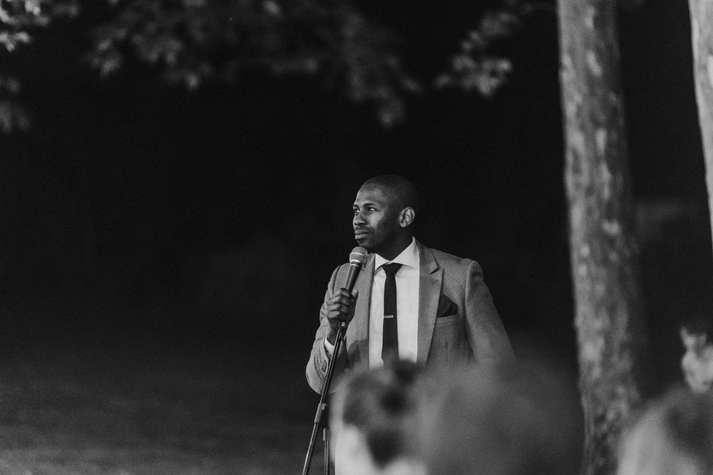 Well-dressed man in suit giving speech at microphone during outdoor evening wedding reception.