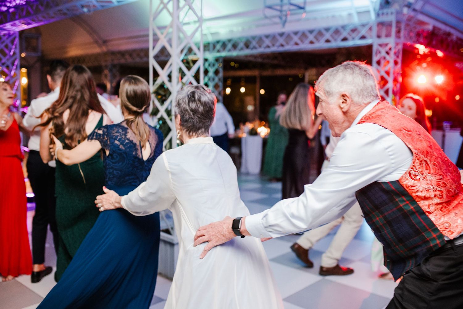 Lively wedding guests dancing together under colorful lights at reception party.
