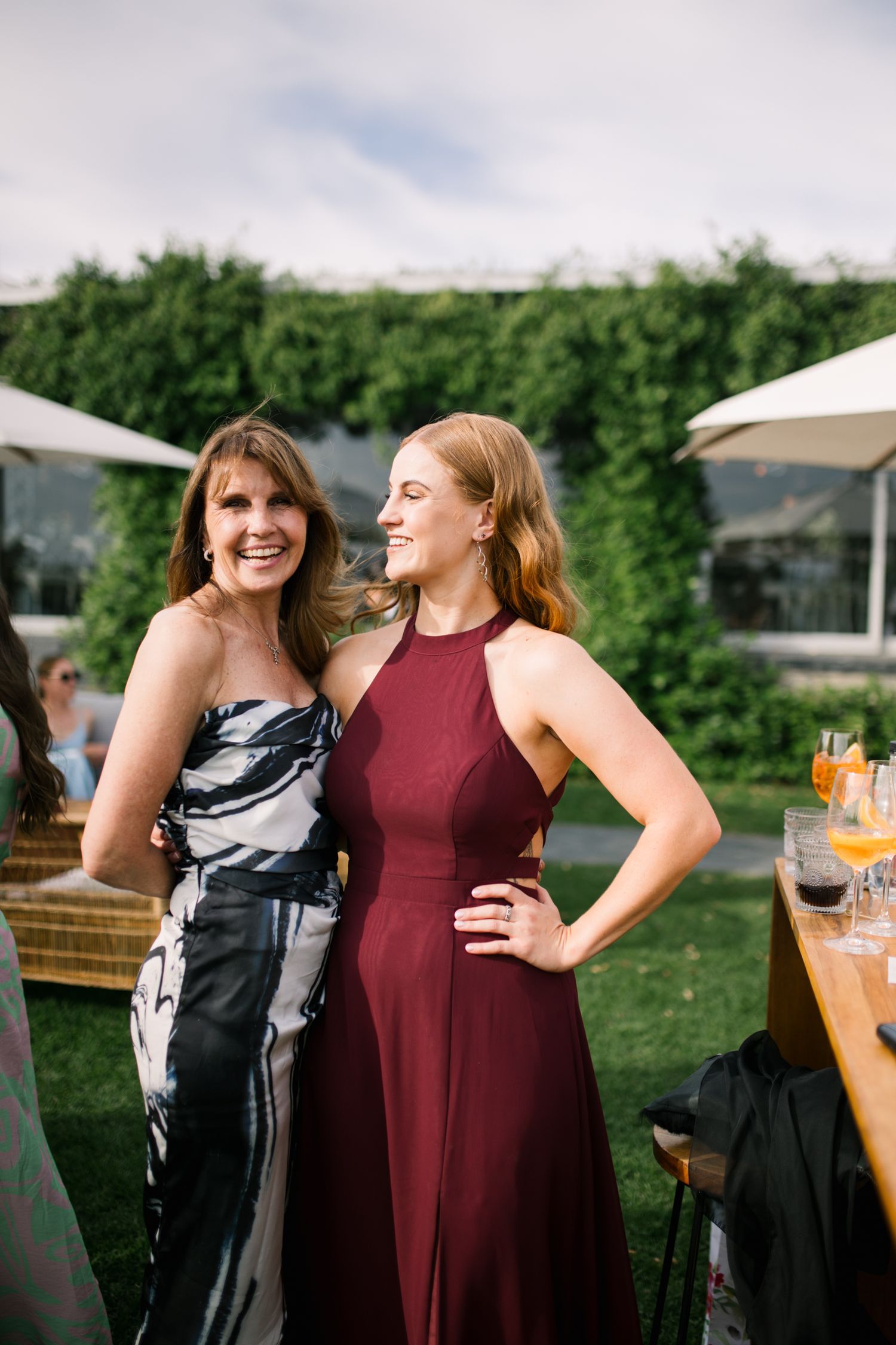 Two smiling female wedding guests in formal gowns posing together at an outdoor garden reception.