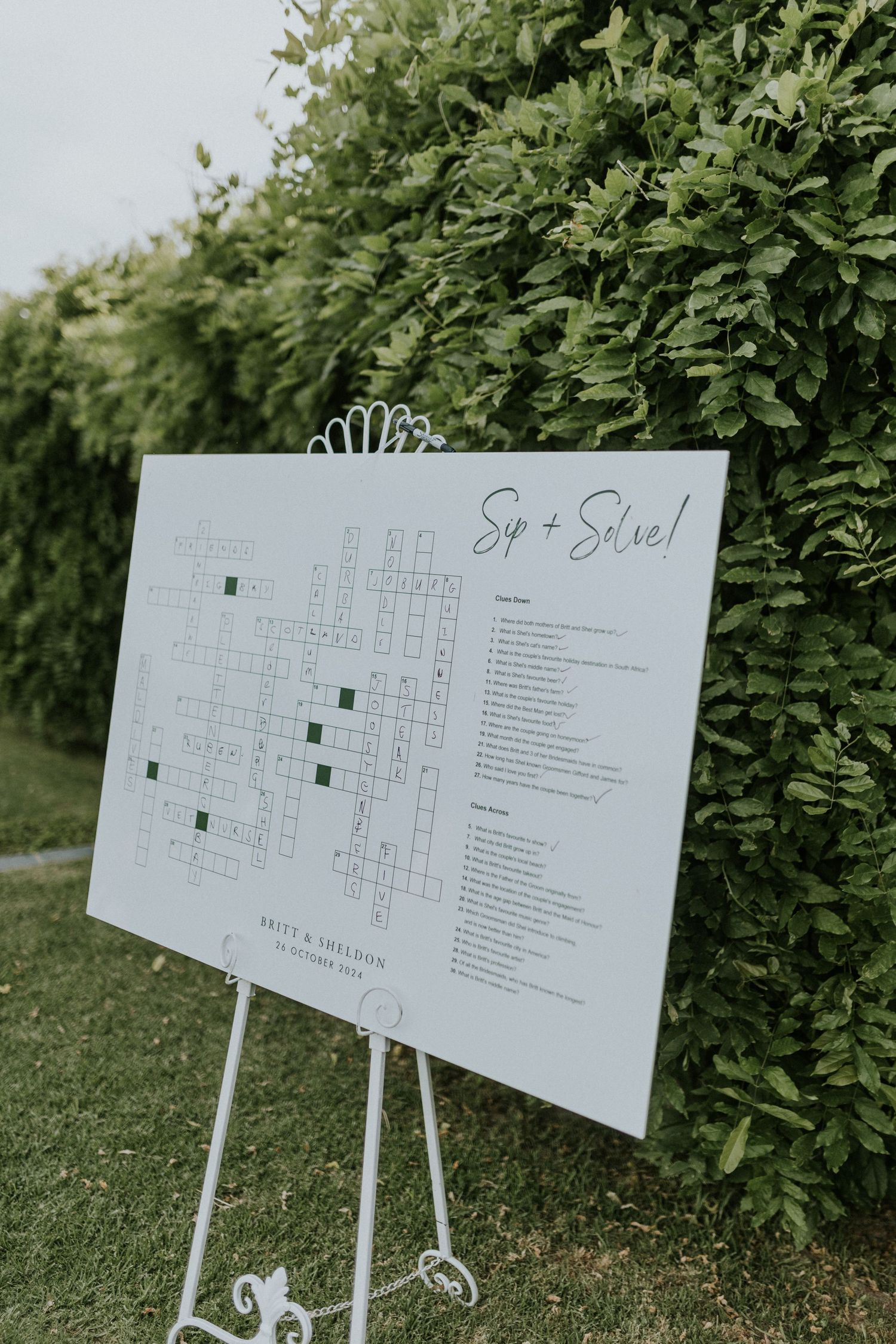 Creative Sip and Solve crossword puzzle seating chart on white easel against lush green hedge backdrop.