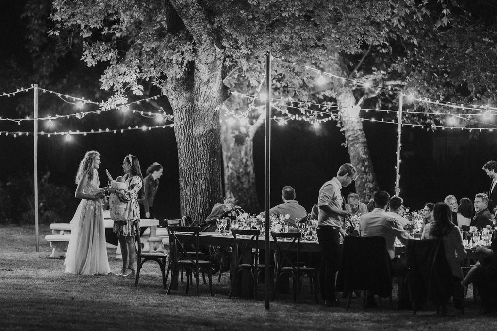 Black and white wide shot of elegant outdoor wedding reception under trees with string lights at night.