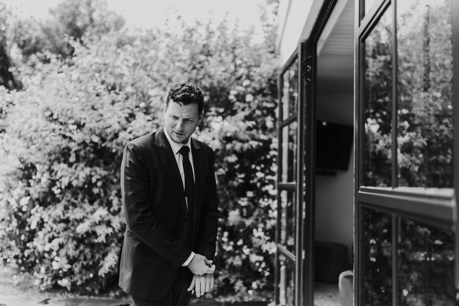 Groom in black suit stands pensively by large windows in black and white photo.