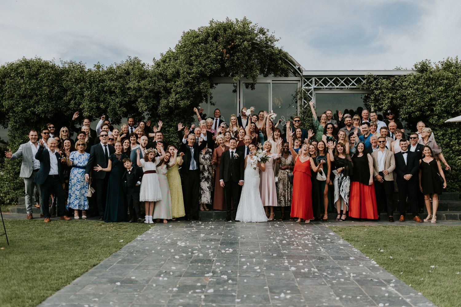 Large wedding party group photo with bride, groom, and all guests celebrating outdoors at an elegant garden venue.