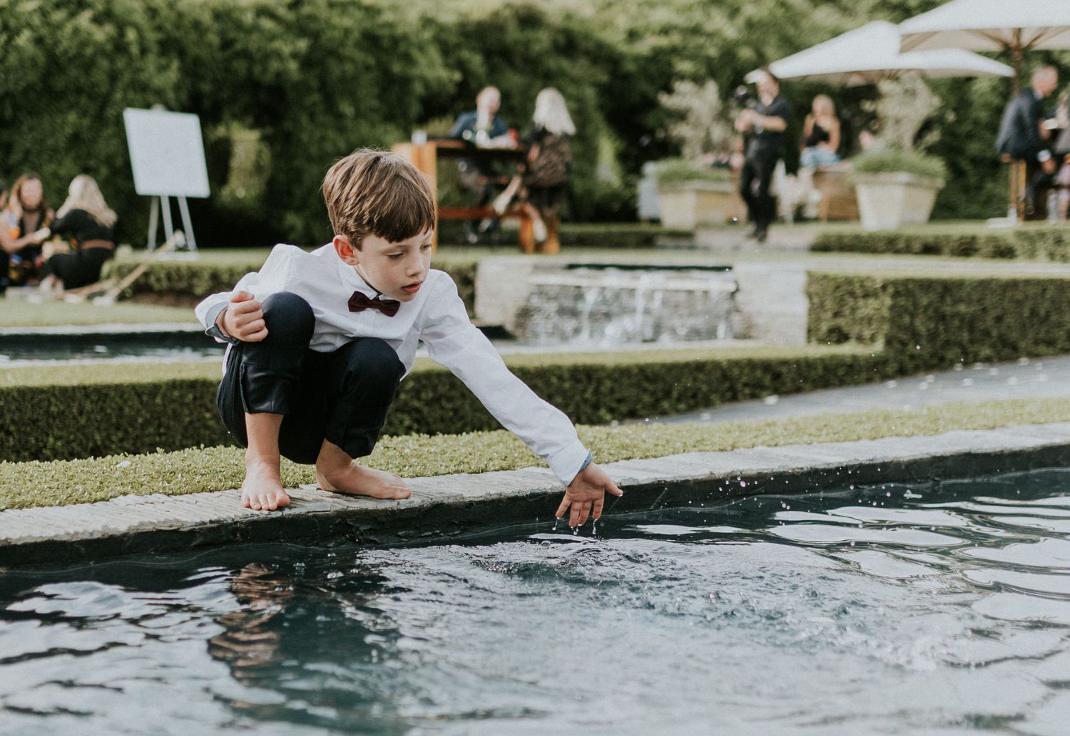 Young boy in white shirt and bow tie playing with water in an ornamental garden fountain at a wedding.