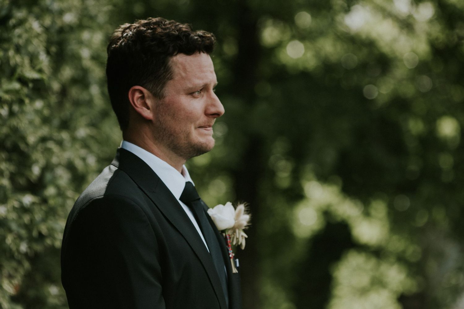 Emotional groom in a dark suit with boutonniere reacts with tears while waiting at the altar surrounded by greenery.