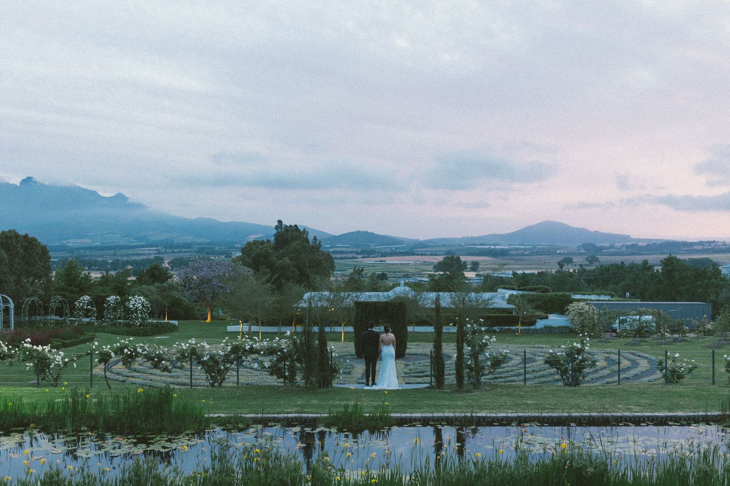 Bride and groom stand together in vast garden estate with stunning mountain backdrop at dusk.