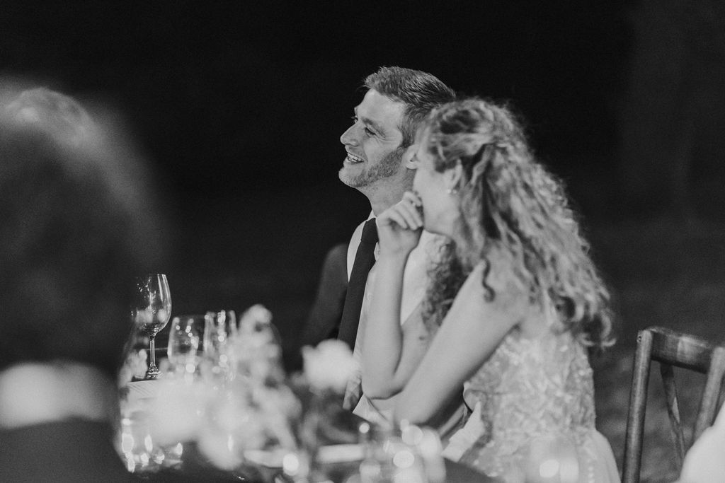 Black and white photo of smiling bride and groom seated at wedding reception dinner table.
