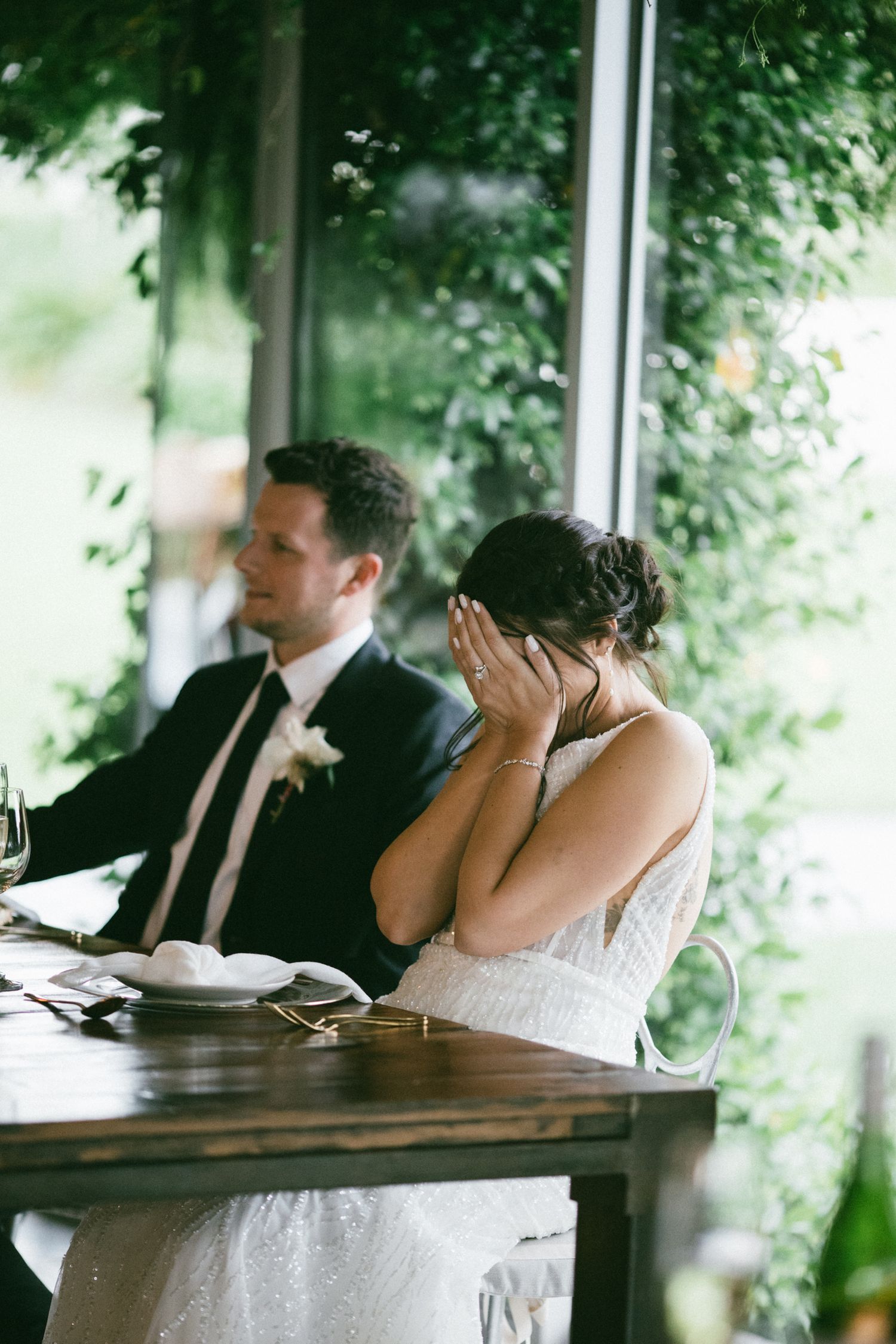 Emotional bride covering her face during wedding speeches while groom sits beside her at reception table.