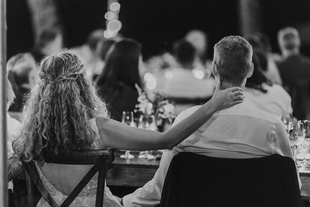 Bride and groom sit together from behind at outdoor wedding reception, groom's arm around bride.