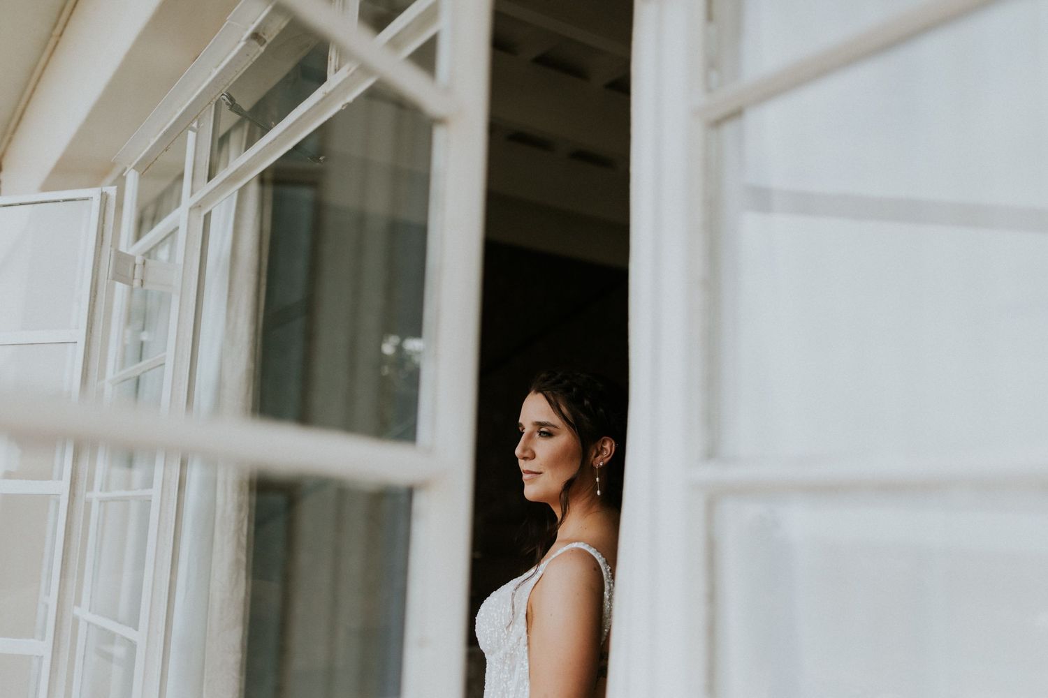 Stunning bride in sleeveless white wedding gown framed by white French doors at venue.