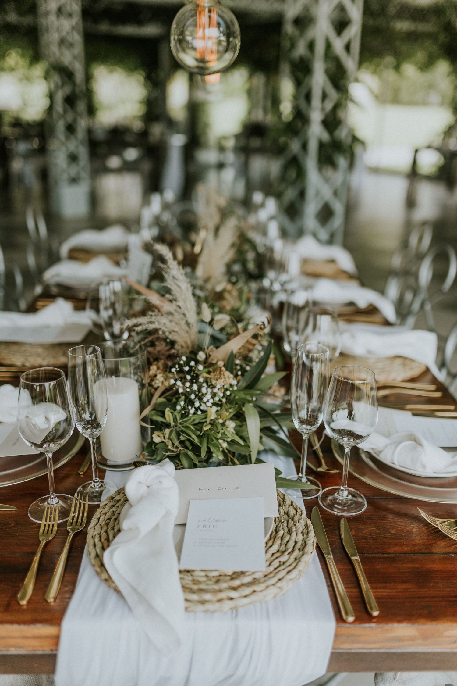 Elegant wedding table setting with woven chargers, gold cutlery, greenery, and candles.