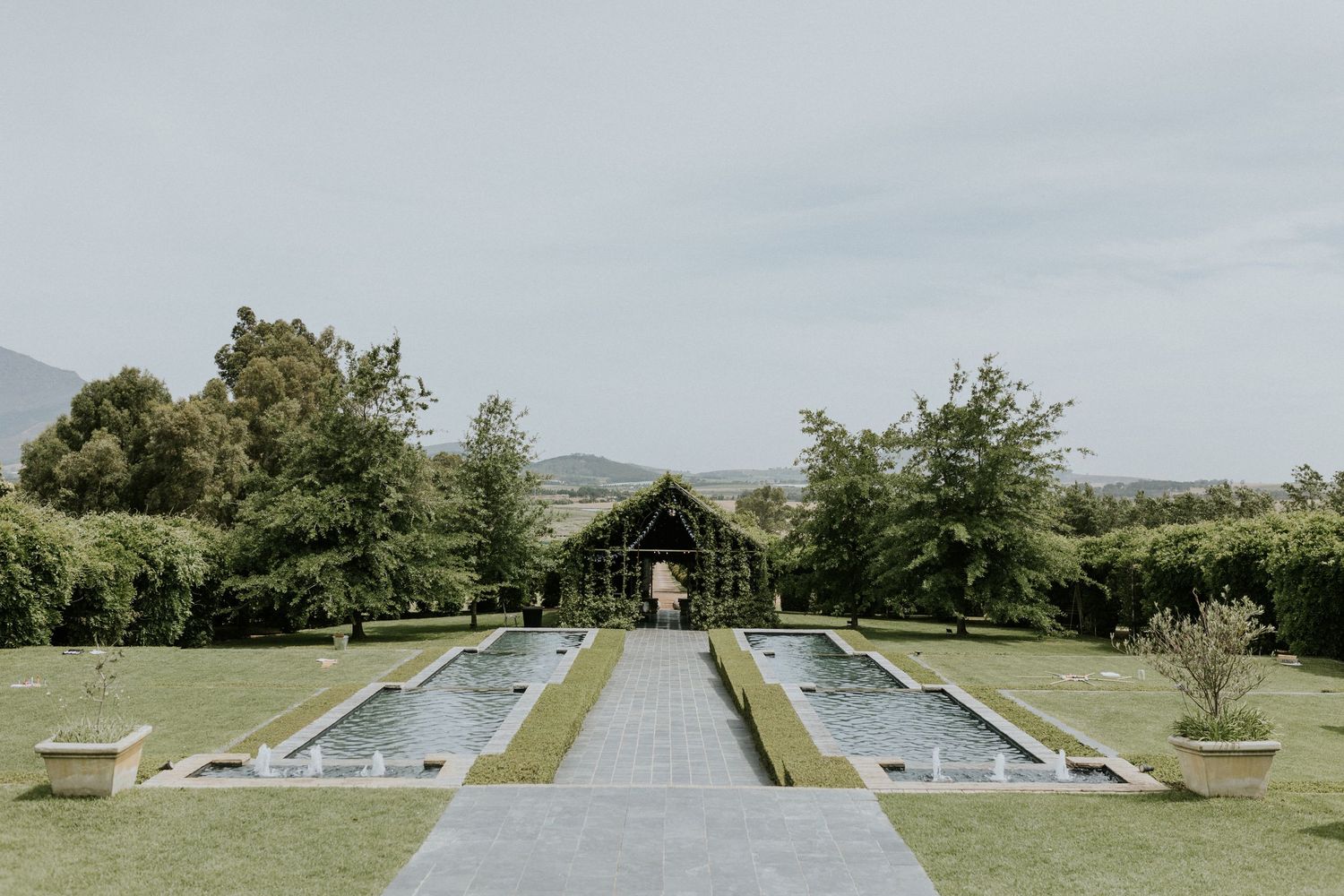 Symmetrical formal garden with twin rectangular reflecting pools, manicured hedges, and ivy-covered pergola with mountain backdrop.