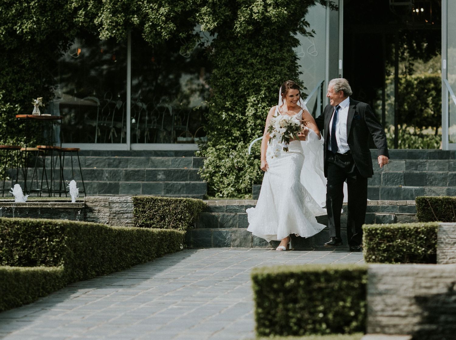 Bride in a white wedding gown walks with her father through a formal garden with manicured hedges before the ceremony.