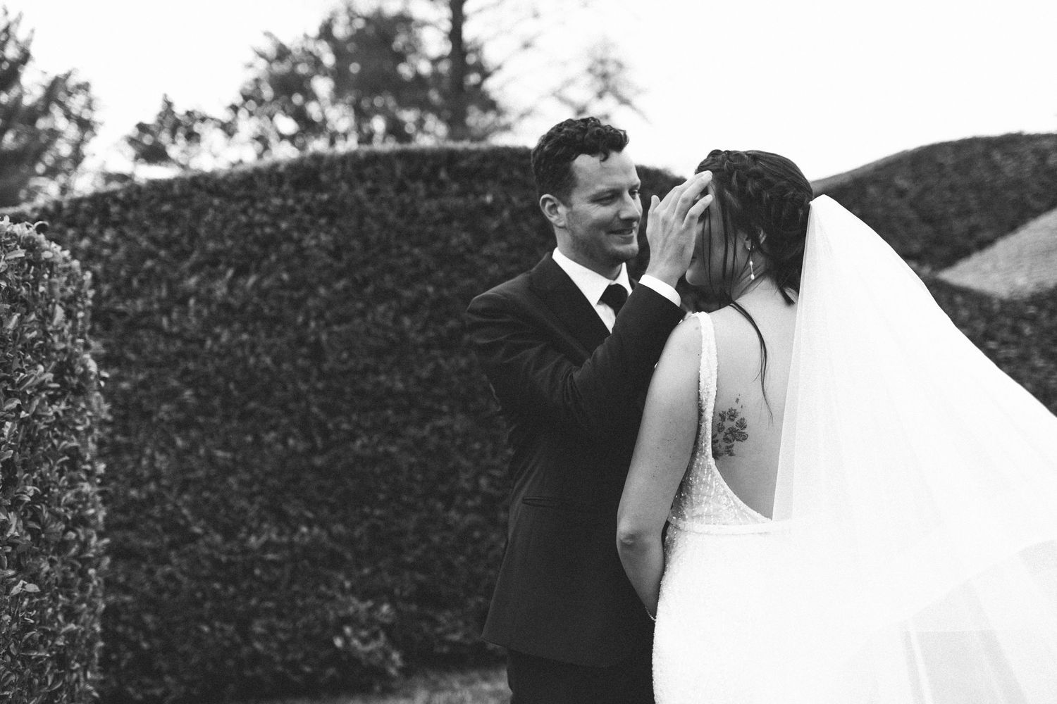 Black and white wedding portrait of groom leaning in to kiss veiled bride outdoors.