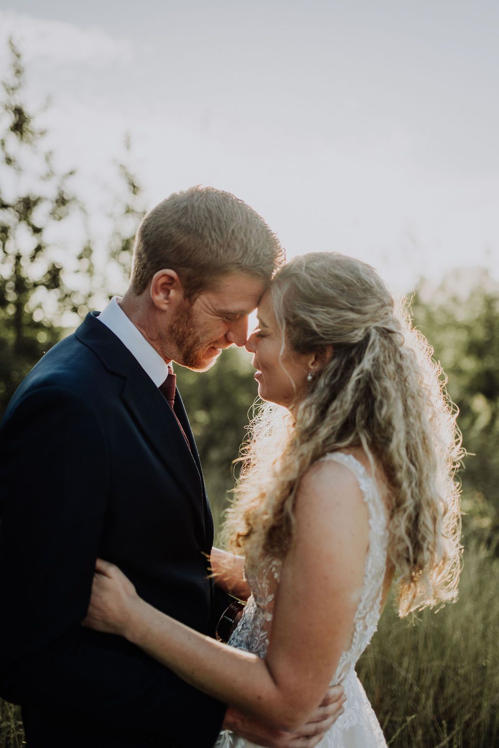 Bride and groom touching foreheads in a tender embrace surrounded by lush greenery at golden hour.