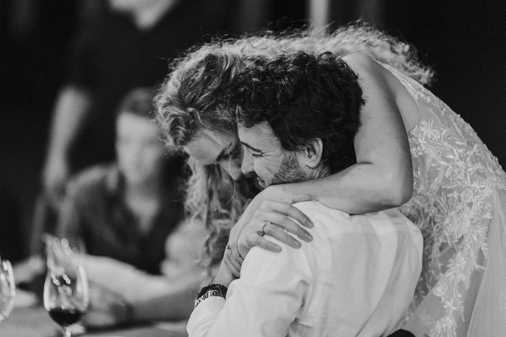 Bride and groom share emotional hug at outdoor wedding reception table.