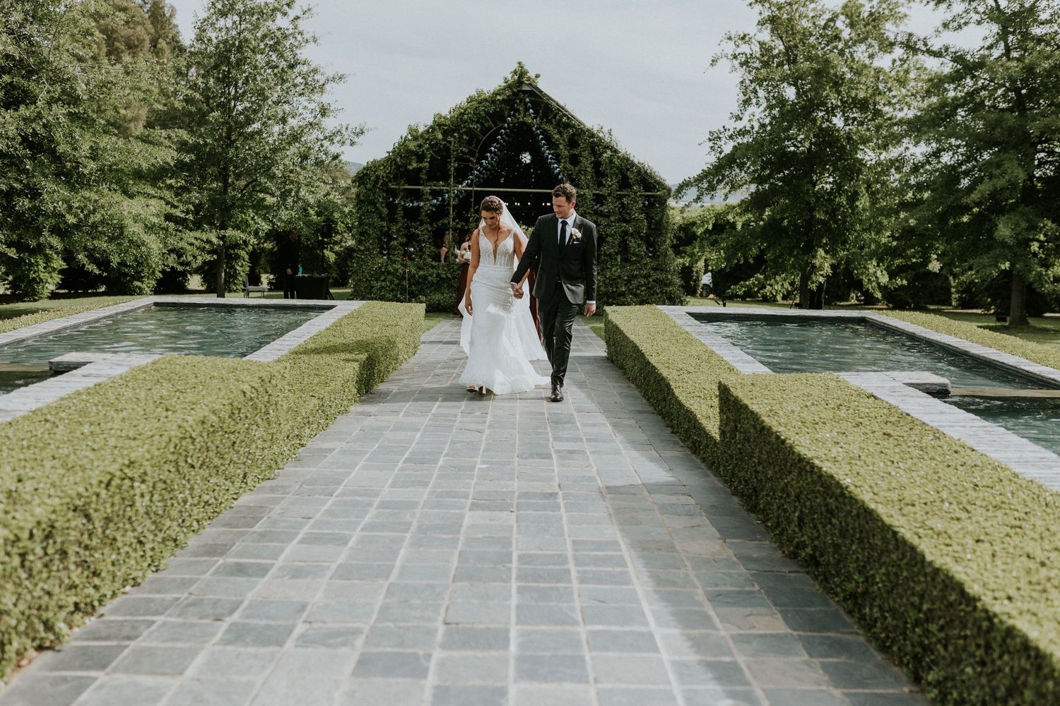 Bride and groom walk hand in hand along a stone pathway surrounded by manicured hedges at an elegant garden wedding venue.