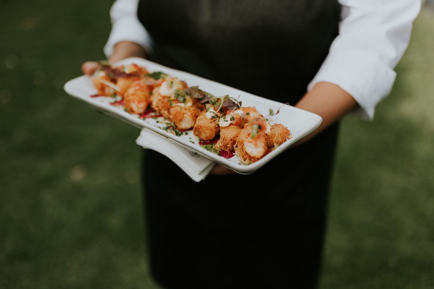 Waiter in black apron serving a platter of golden fried shrimp appetizers at an outdoor event.