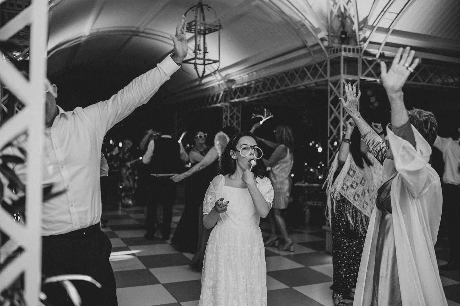 Bride singing into microphone with hands raised on checkered dance floor at lively wedding reception.
