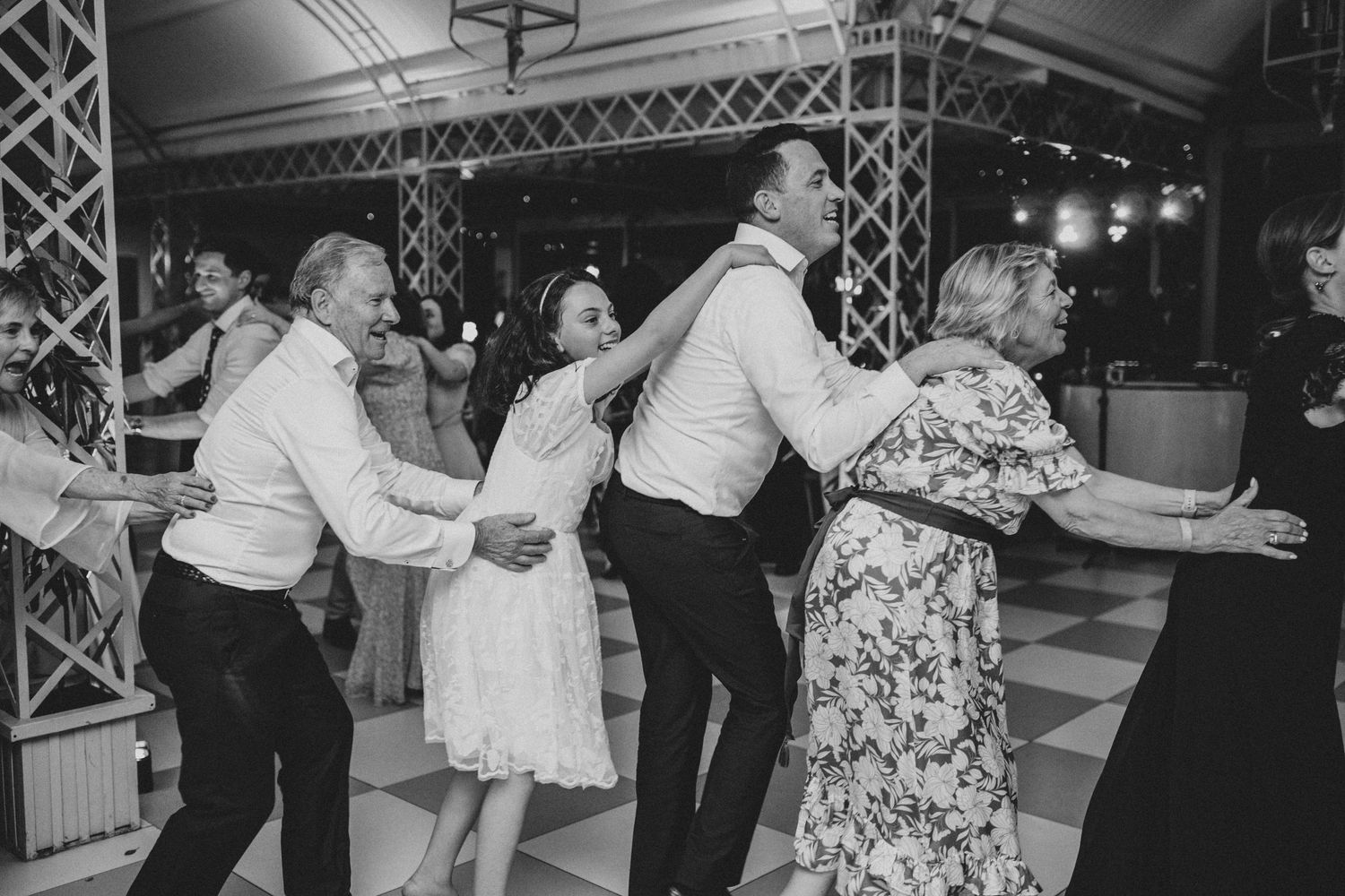 Wedding guests dancing together in a lively conga line on a checkered dance floor at a reception.